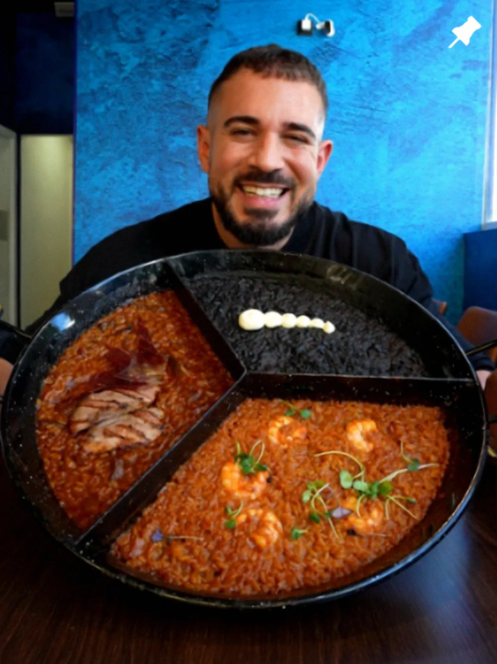 El cocinero sostiene la paellera del trío de arroces delante de la pared azul del restaurante